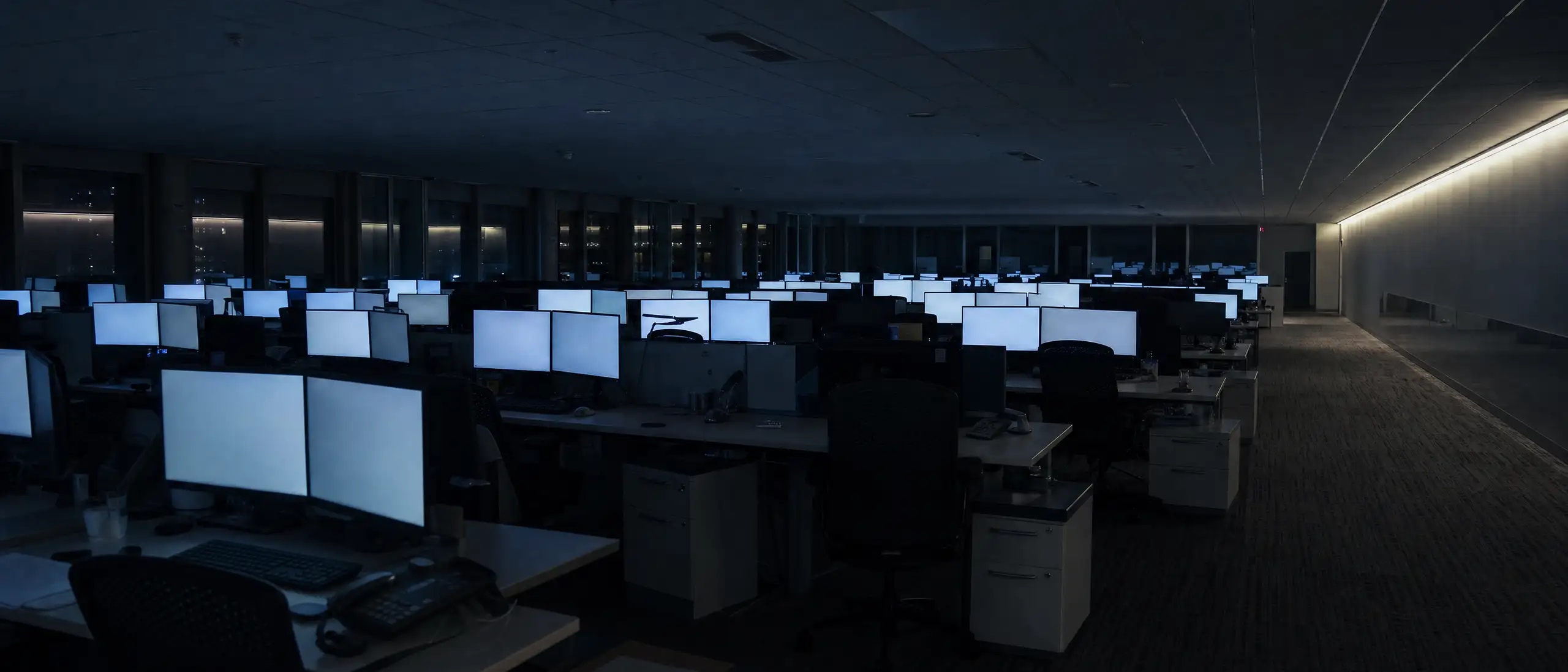 A large open-plan office floor at night. Rows of empty desks with monitors still switched on, overhead lights off, a lit strip along the far wall.